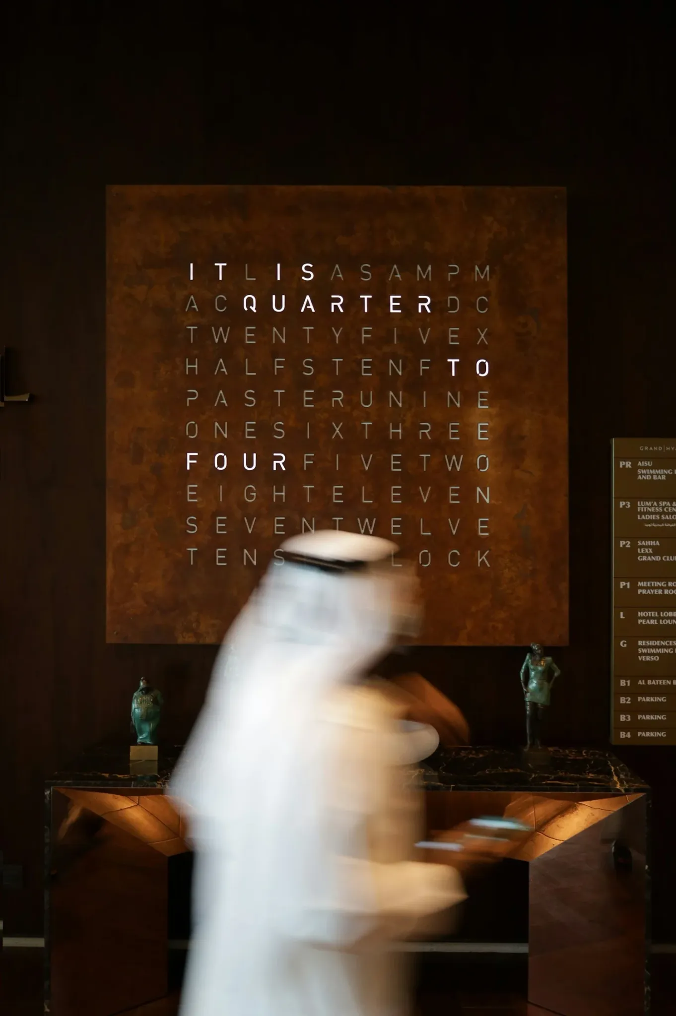A blurred figure passing a sculptural word clock in a Gulf interior